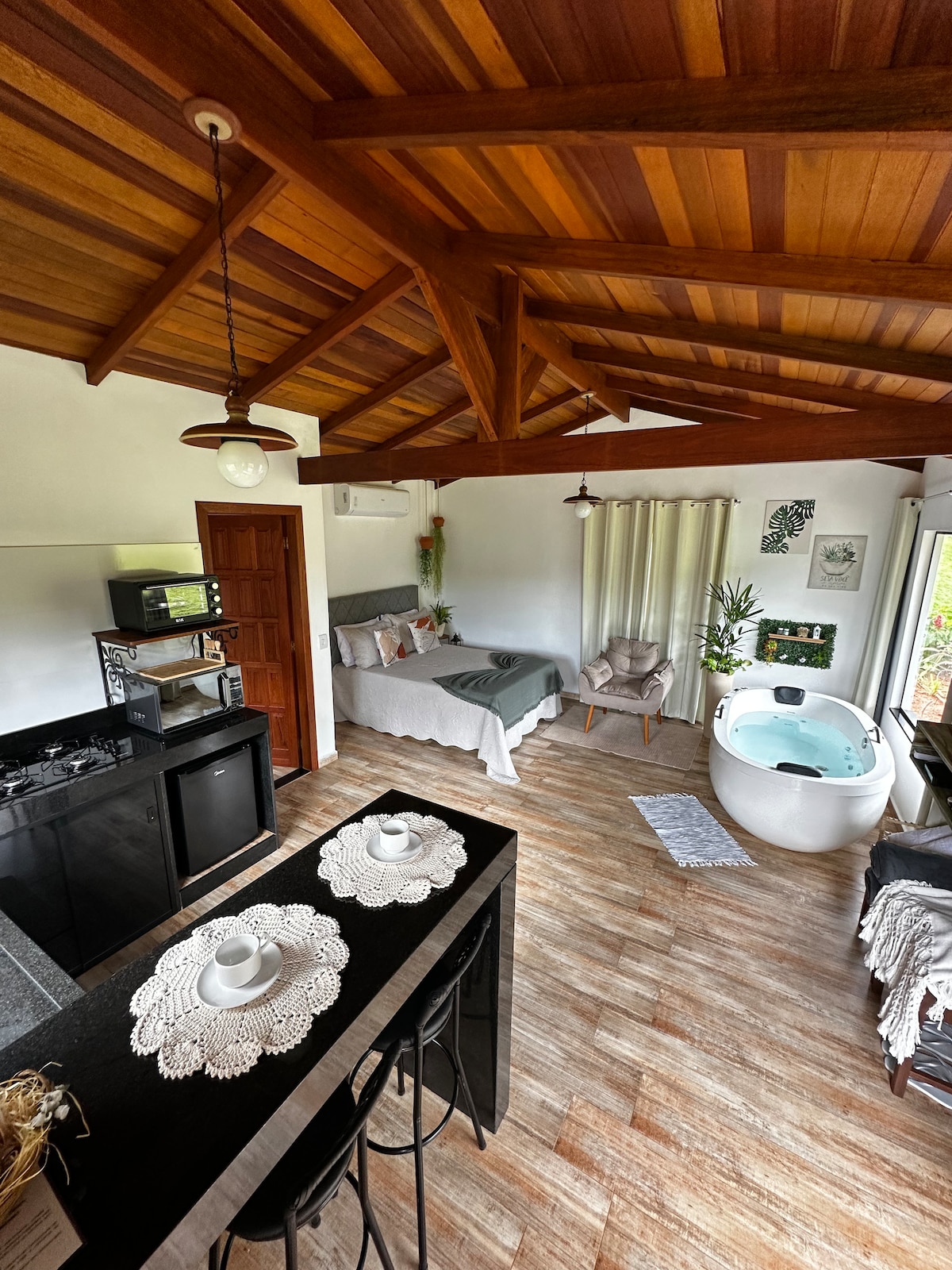 An open living space features a well-equipped kitchen area, a spacious bed adorned with soft linens, and a unique freestanding bathtub. Large windows allow natural light to fill the room, highlighting the warm wooden ceiling beams and inviting décor.