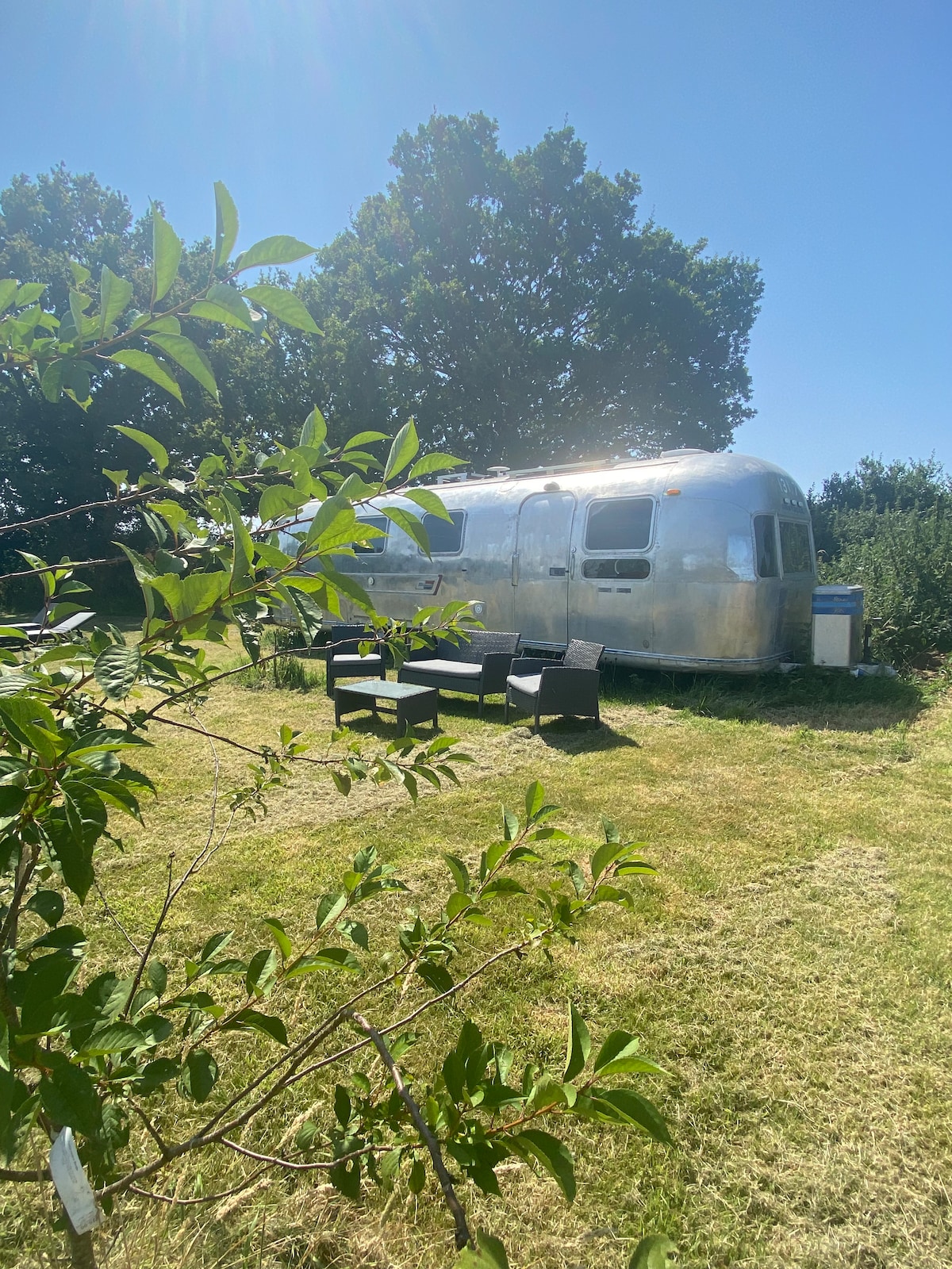 The Airstream Sovereign is nestled in a grassy area surrounded by lush greenery. A seating arrangement of dark wicker chairs is placed nearby, offering a spot to relax outdoors. Sunlight glints off the metallic exterior, enhancing the tranquil ambiance of the countryside.