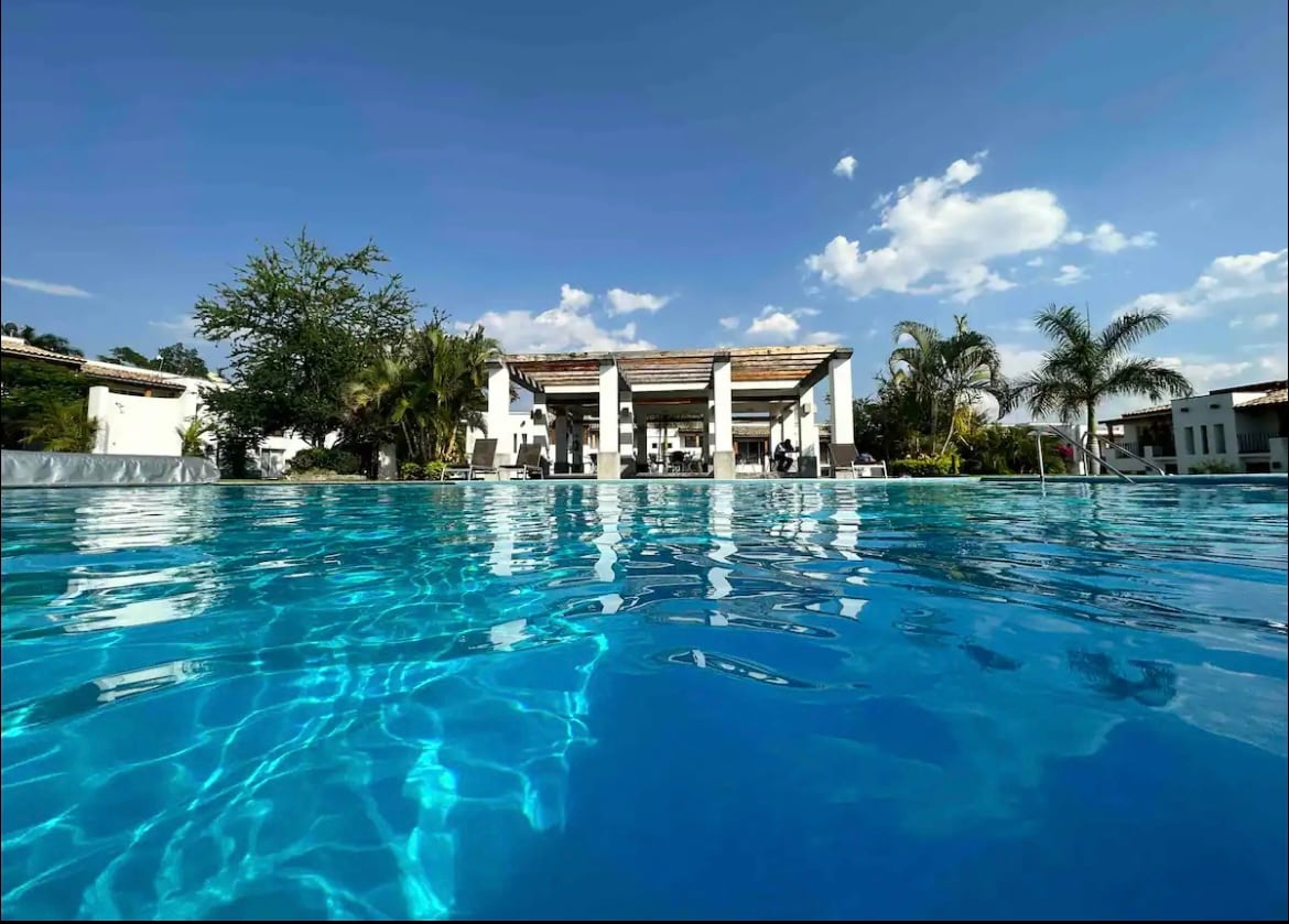 A clear view of the picturesque pool reflects the bright sky above. The backdrop features a spacious outdoor area with palm trees and a deck, inviting relaxation. Lounge chairs are positioned under a pergola, offering shaded seating near the water.