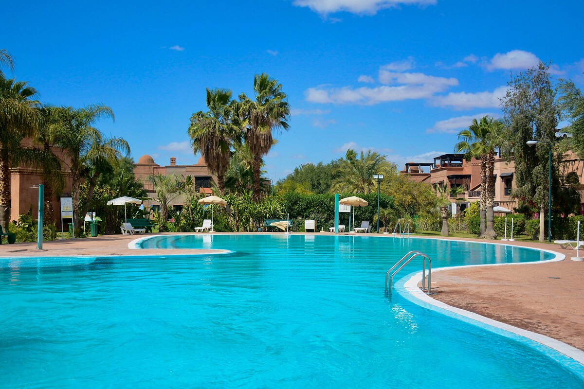 A large, inviting swimming pool features crystal-clear blue water surrounded by lush palm trees and vibrant greenery. Sun loungers and umbrellas are positioned along the pool's edge, creating a relaxing atmosphere for guests to enjoy.