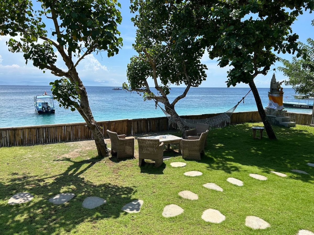 A grassy outdoor area features two trees providing shade, with a seating arrangement made of natural materials positioned on a stone pathway. A view of the ocean and a boat can be seen in the distance, enhancing the coastal environment.