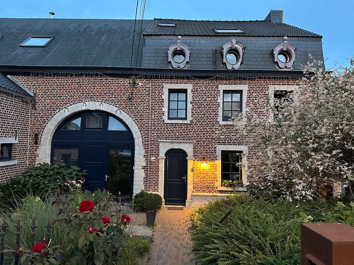 The exterior of a historic brick building is showcased, featuring arched doorways and decorative dormer windows. Lush greenery and flowering plants frame the entrance, adding a touch of color and life. Soft lighting highlights the doorway, creating an inviting appearance during twilight.