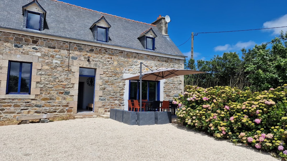 The exterior of a stone house features a light gray facade with blue window frames. A patio area with an umbrella is visible, surrounded by a landscaped yard with blooming hydrangeas. A gravel pathway leads to the entrance, enhancing the inviting atmosphere.
