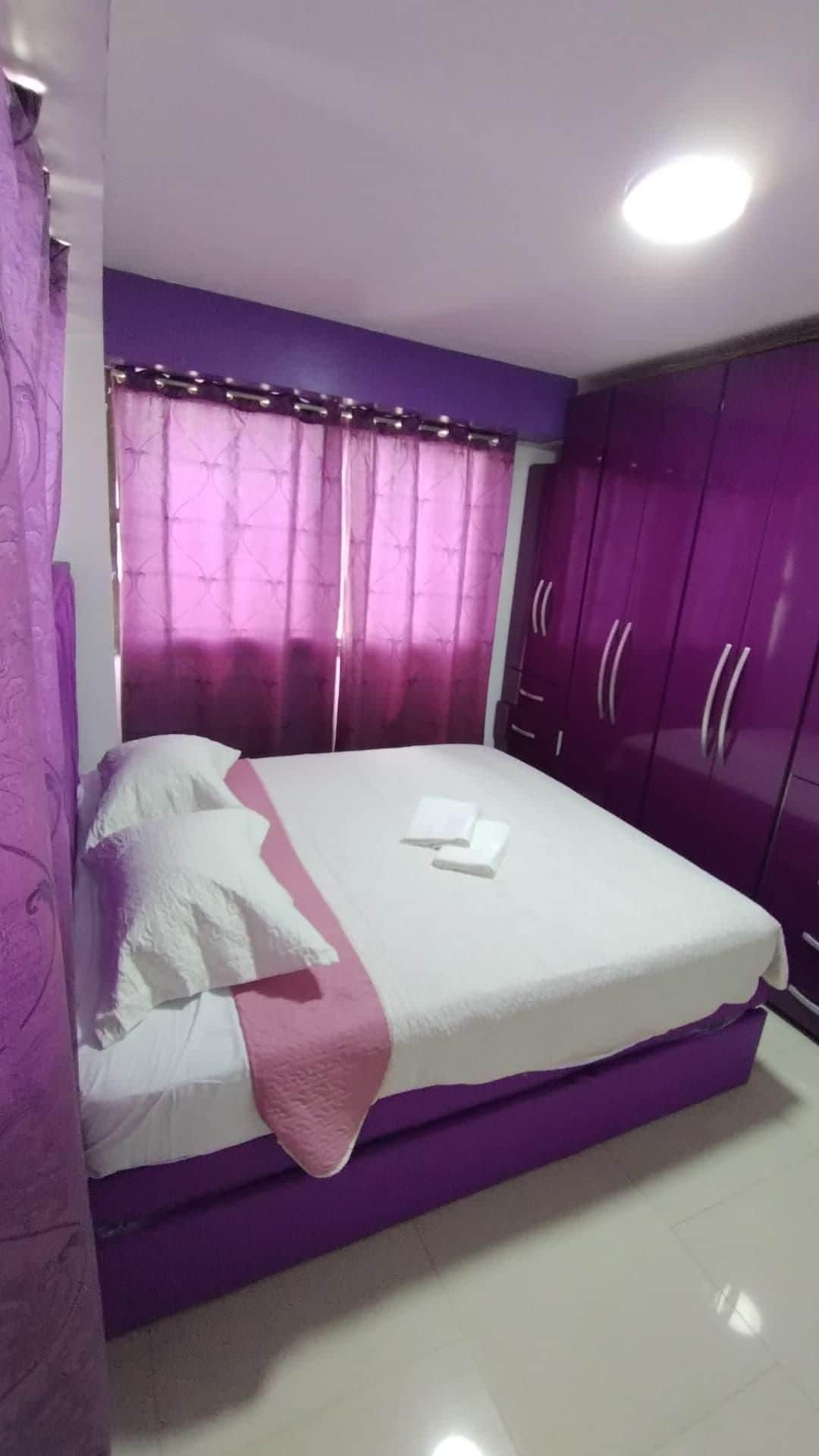 A modern bedroom features a spacious bed dressed in white linens with a light pink throw. Rich purple curtains drape over the window, complemented by a large purple wardrobe that offers ample storage. Soft lighting enhances the welcoming ambiance of the space.