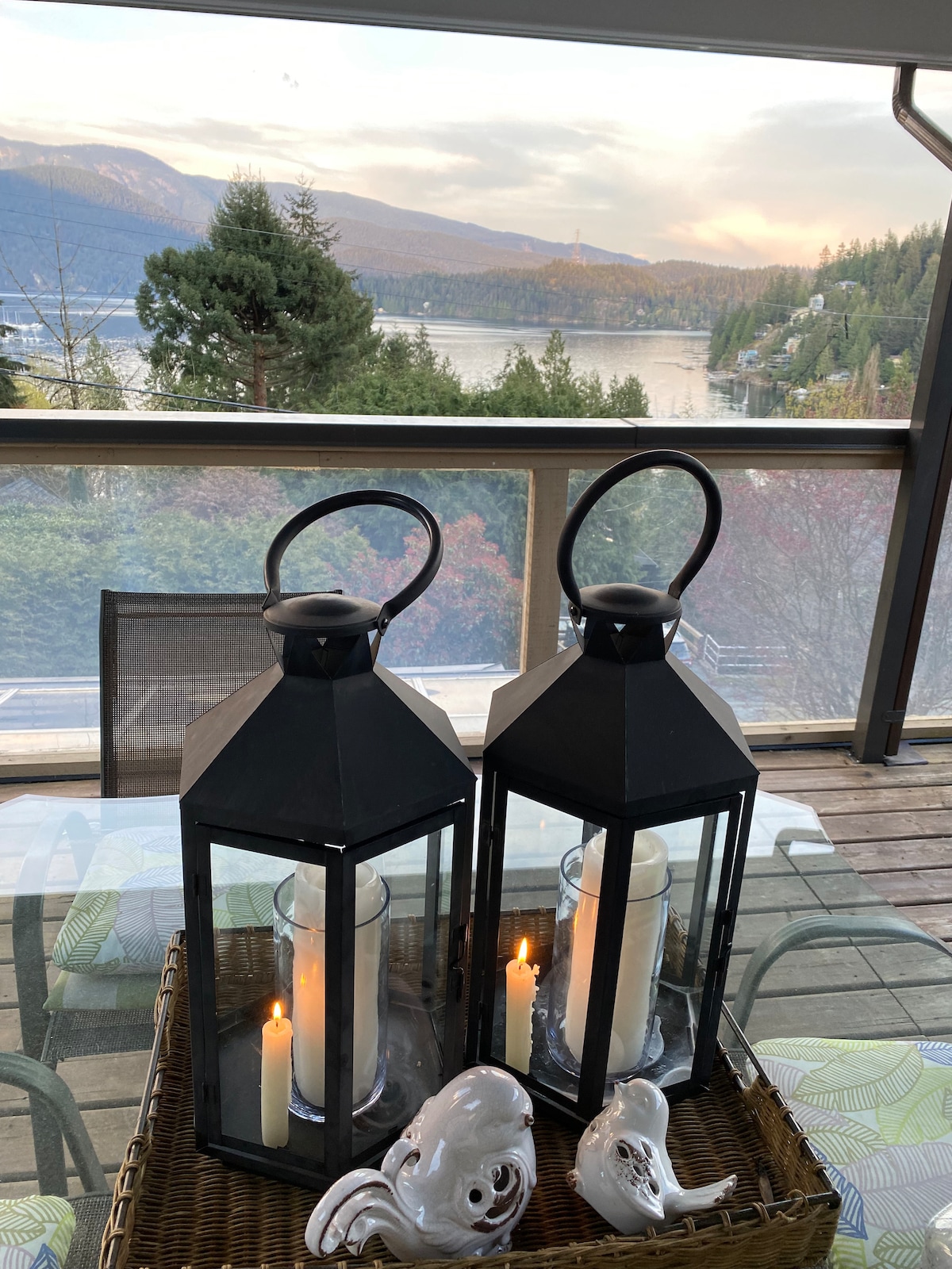 Two black lanterns are positioned on a wicker tray, each containing a candle. The background showcases a scenic view of the mountains and water, with soft natural lighting enhancing the landscape. A glimpse of outdoor seating is visible, framed by the deck.
