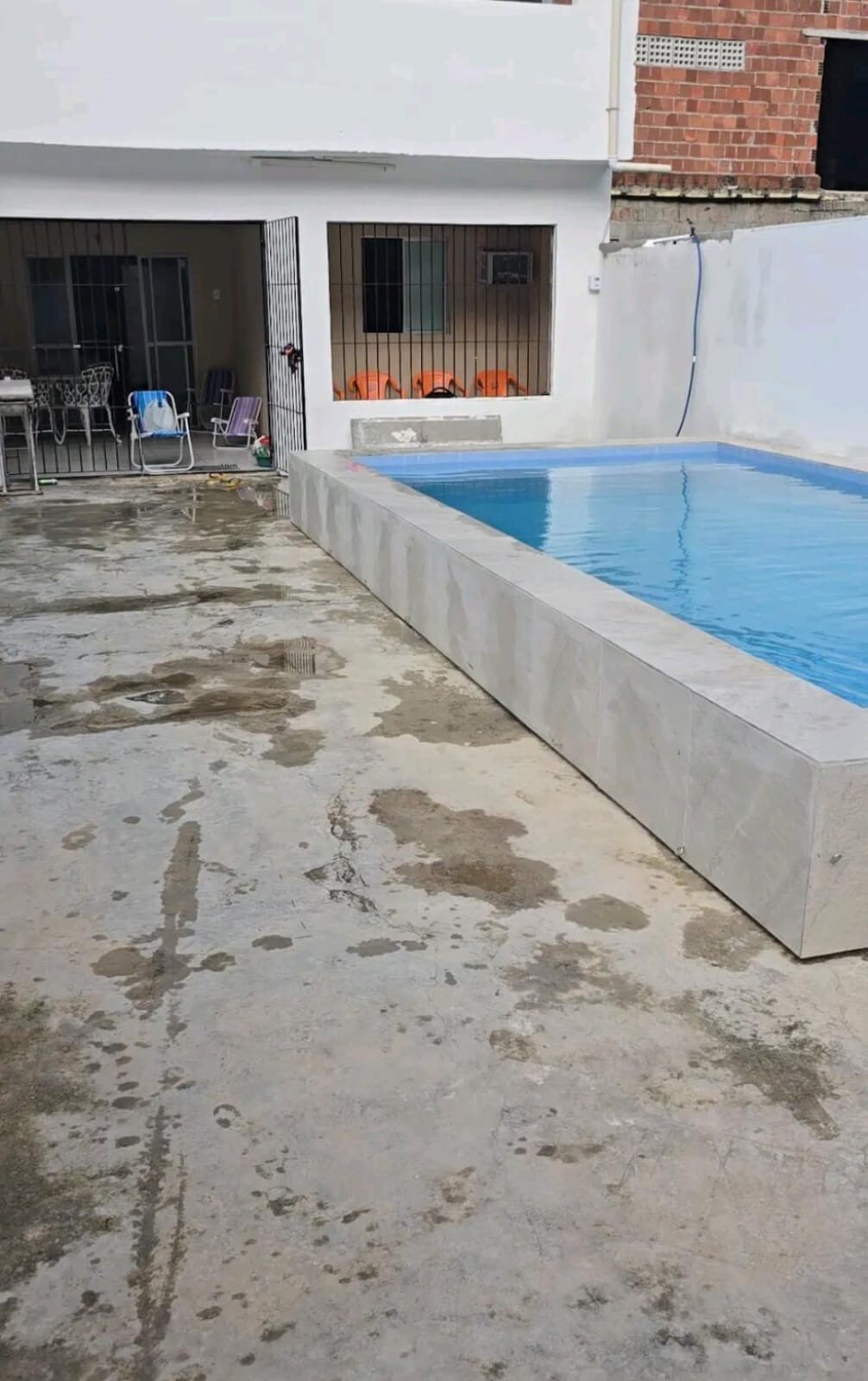 A clean and modern outdoor space features a rectangular swimming pool with clear blue water, surrounded by a smooth concrete deck. In the background, a sliding door leads to a living area with outdoor seating visible.