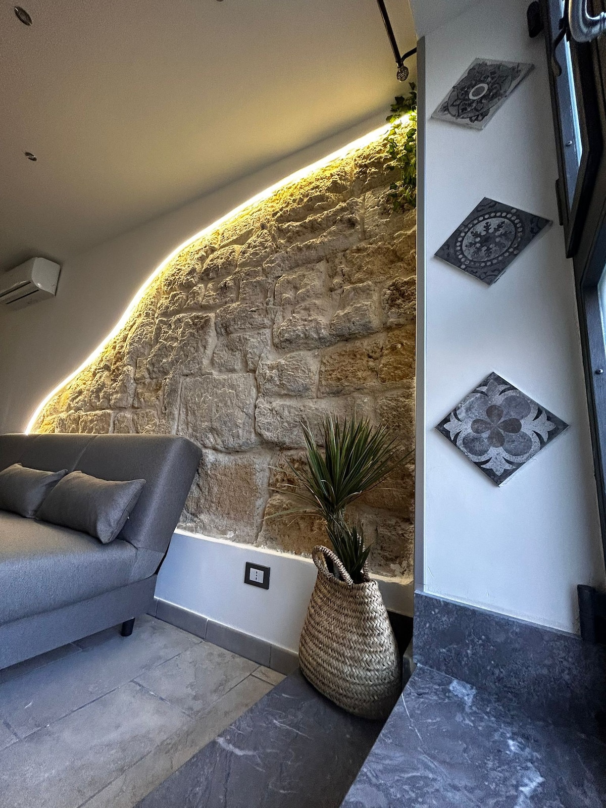 A cozy corner of the living area features a gray sofa with decorative cushions, complemented by an illuminated stone wall. A potted plant sits in a woven basket, while wall decorations add subtle character to the space.