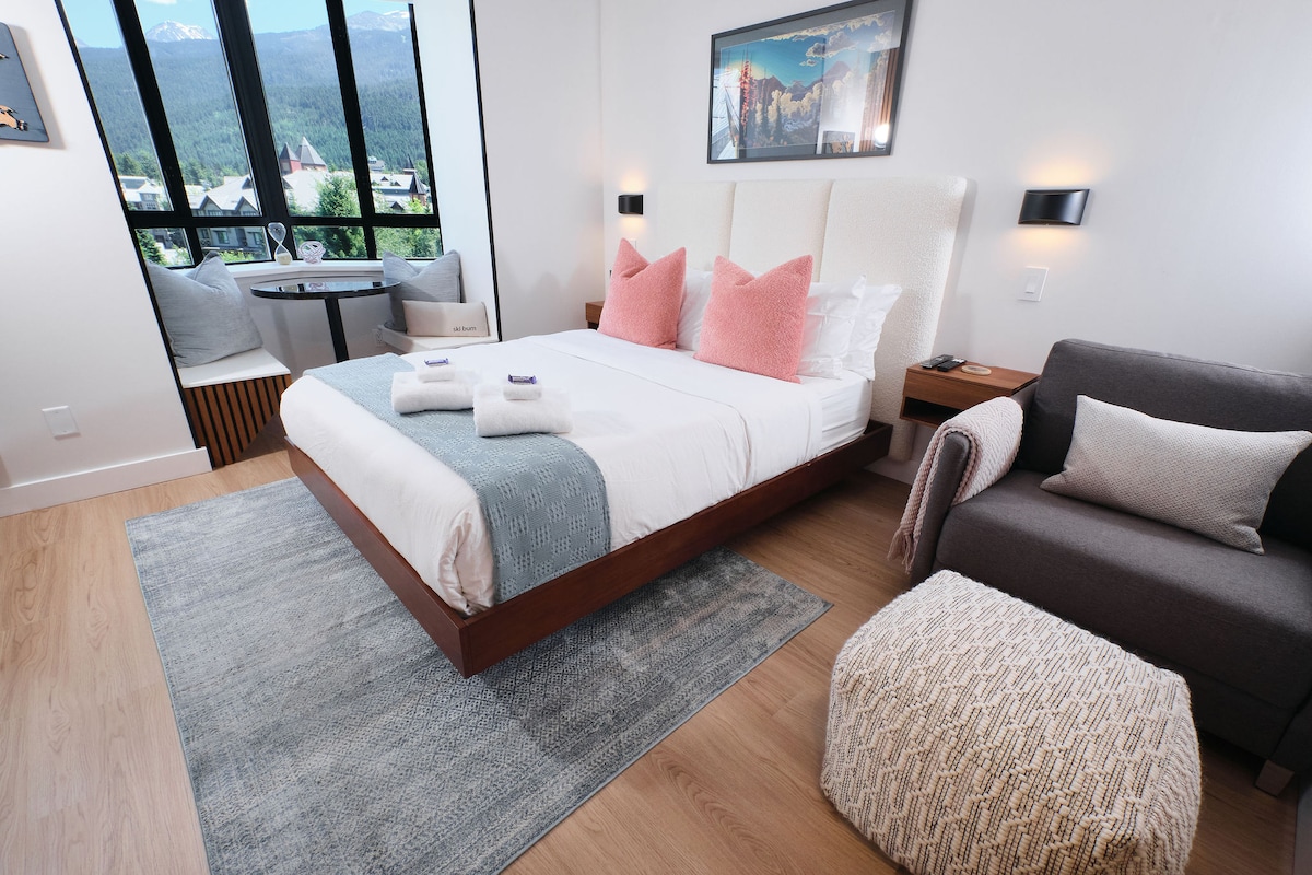 A spacious studio features a queen-size bed with decorative pillows and a cozy designer chair. Natural light floods the room from large windows, offering mountain views. A soft rug lies beneath the bed, while a small seating area adds functionality.