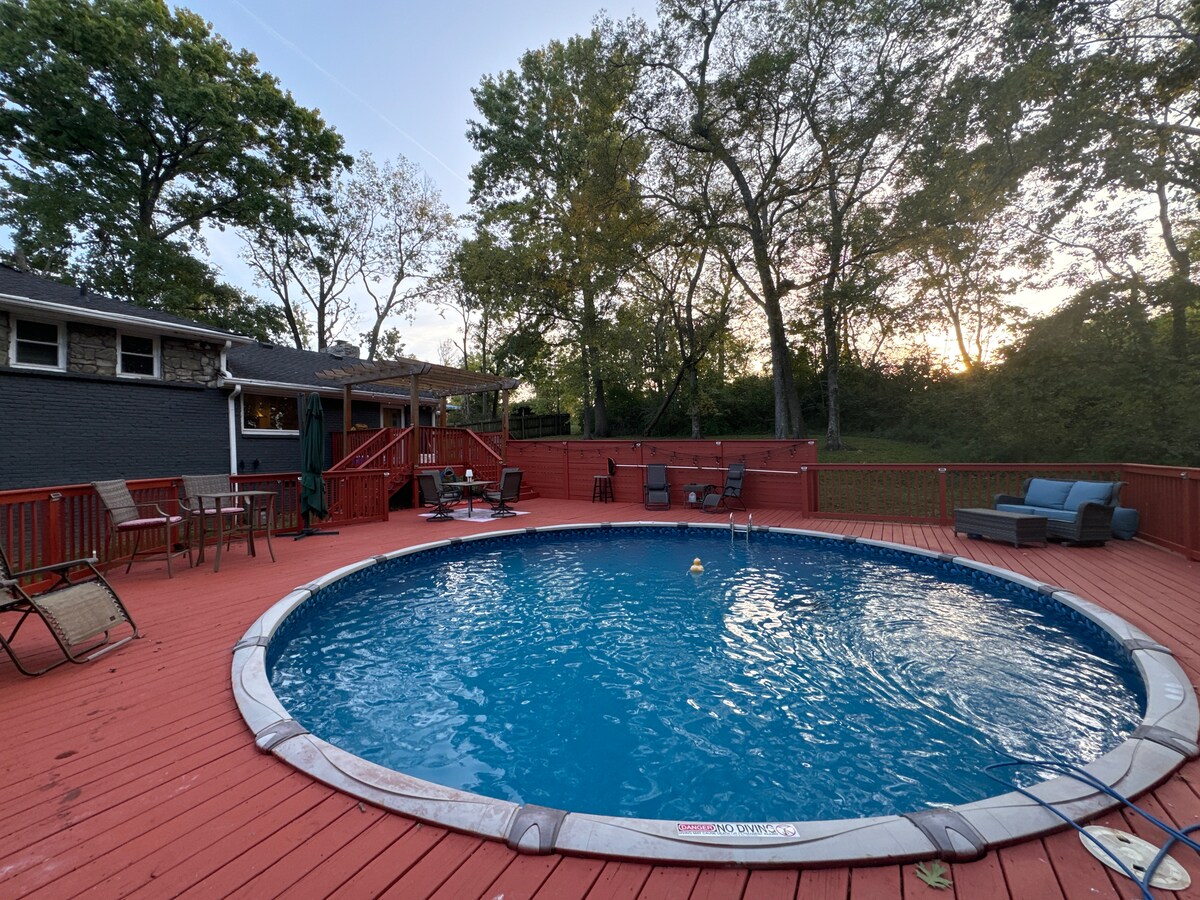 A circular pool is surrounded by a spacious wooden deck, featuring lounge chairs and a section with seating options. Trees in the background frame the area, while a soft glow of sunset adds a serene ambiance. The pool is currently closed until March.