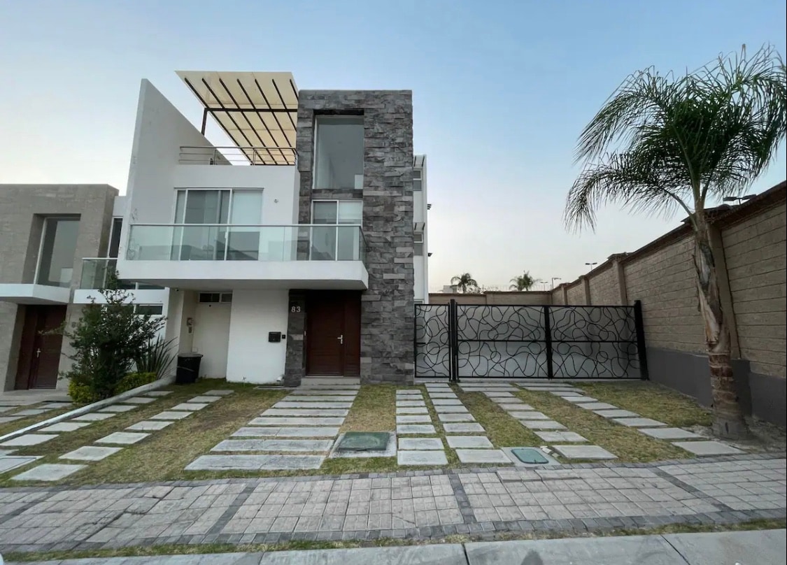 A modern two-story house is presented, featuring a stone accent wall and a balcony with a sleek railing. The front yard showcases a manicured lawn with stepping stones leading to the entrance. A palm tree adds a touch of greenery beside the driveway.