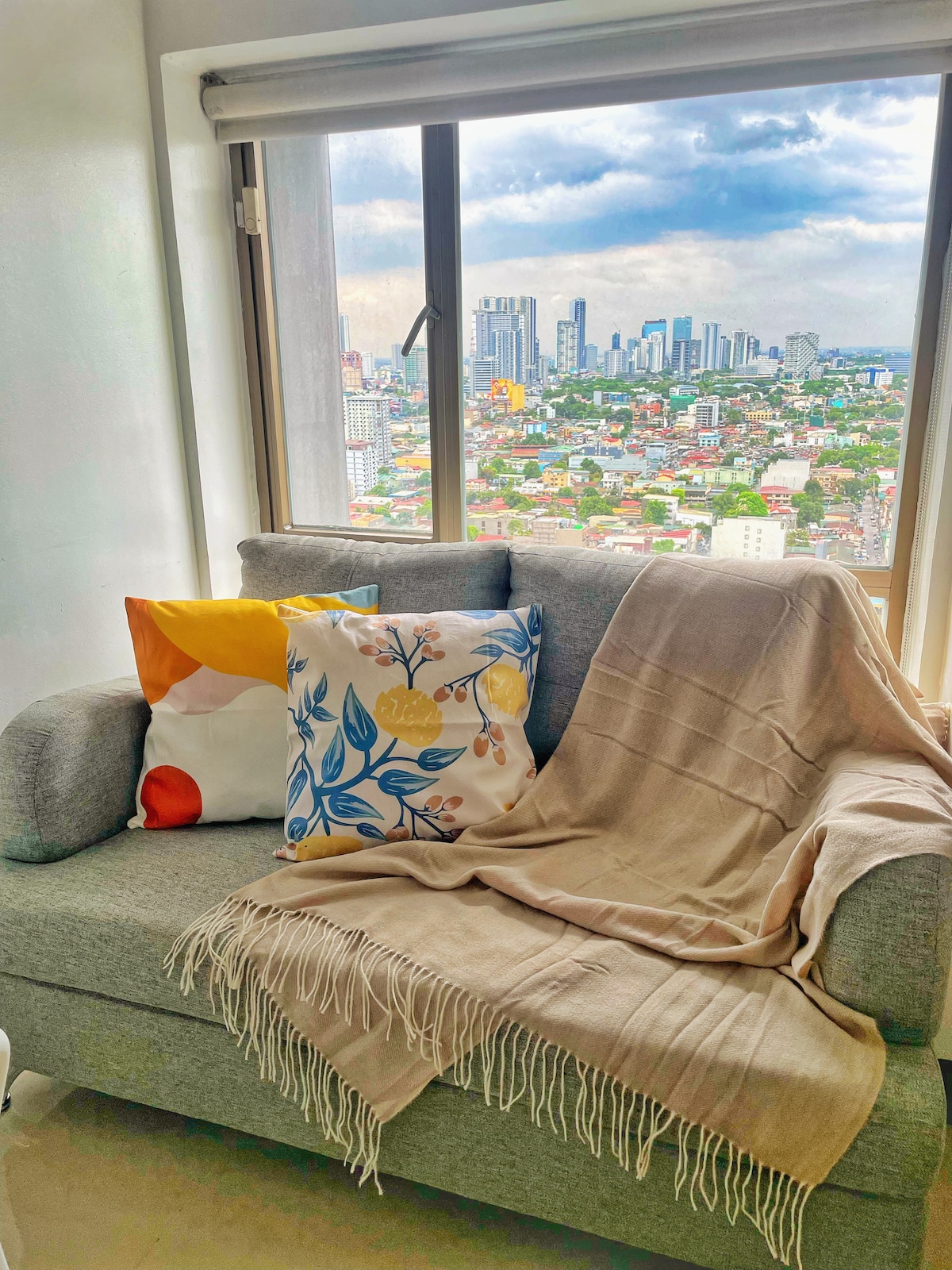 A cozy seating area is presented with a gray sofa adorned with colorful throw pillows. A soft beige blanket is draped over the armrest. Large windows offer a panoramic view of the city skyline, with its diverse buildings and lush rooftops under a partly cloudy sky.