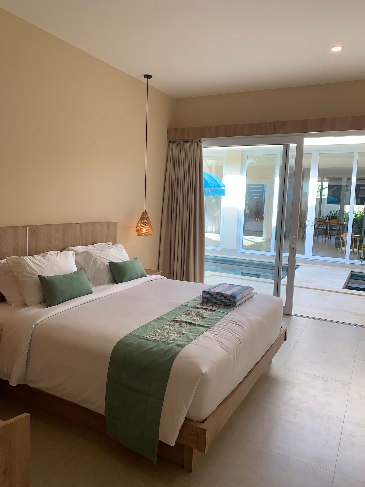 A cozy bedroom features a spacious bed with neatly arranged pillows, complemented by green accents. Natural light streams in through sliding glass doors, leading to an outdoor area. A decorative pendant light hangs above the bed, enhancing the serene environment.