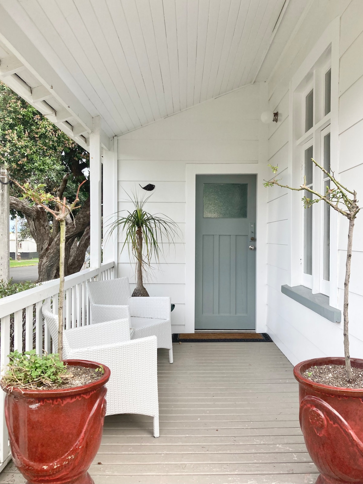 A welcoming porch area is seen, featuring a light grey door at the entrance. Two white wicker chairs are positioned beside small potted plants. Stylish red planters add a pop of color, while the wooden flooring is neatly maintained, creating an inviting entrance.