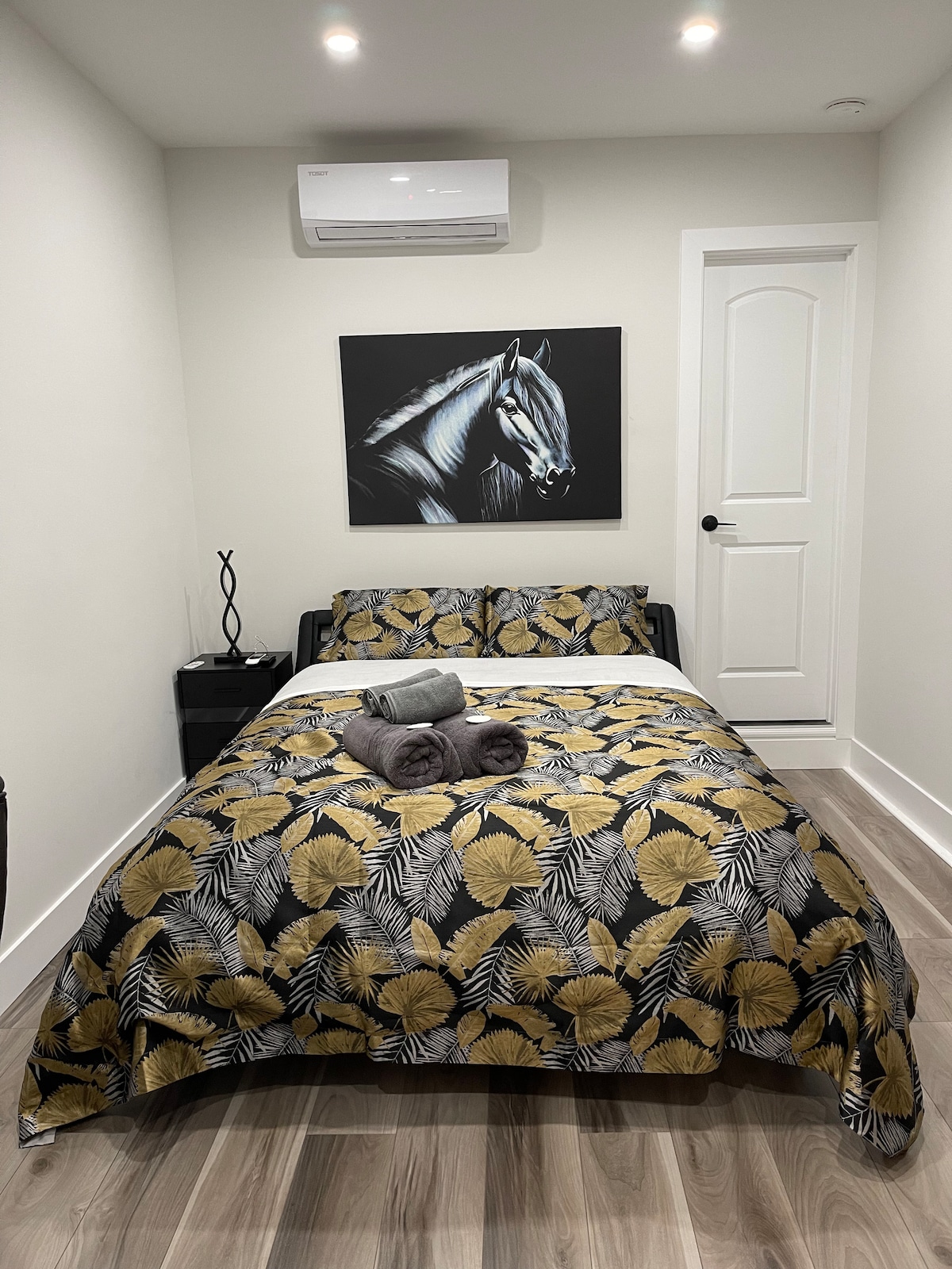A well-appointed room features a queen bed dressed in a patterned comforter with shades of yellow and black. Decorative towels are neatly arranged on the bed. A modern artwork of a horse hangs above the bed, while soft lighting illuminates the space.
