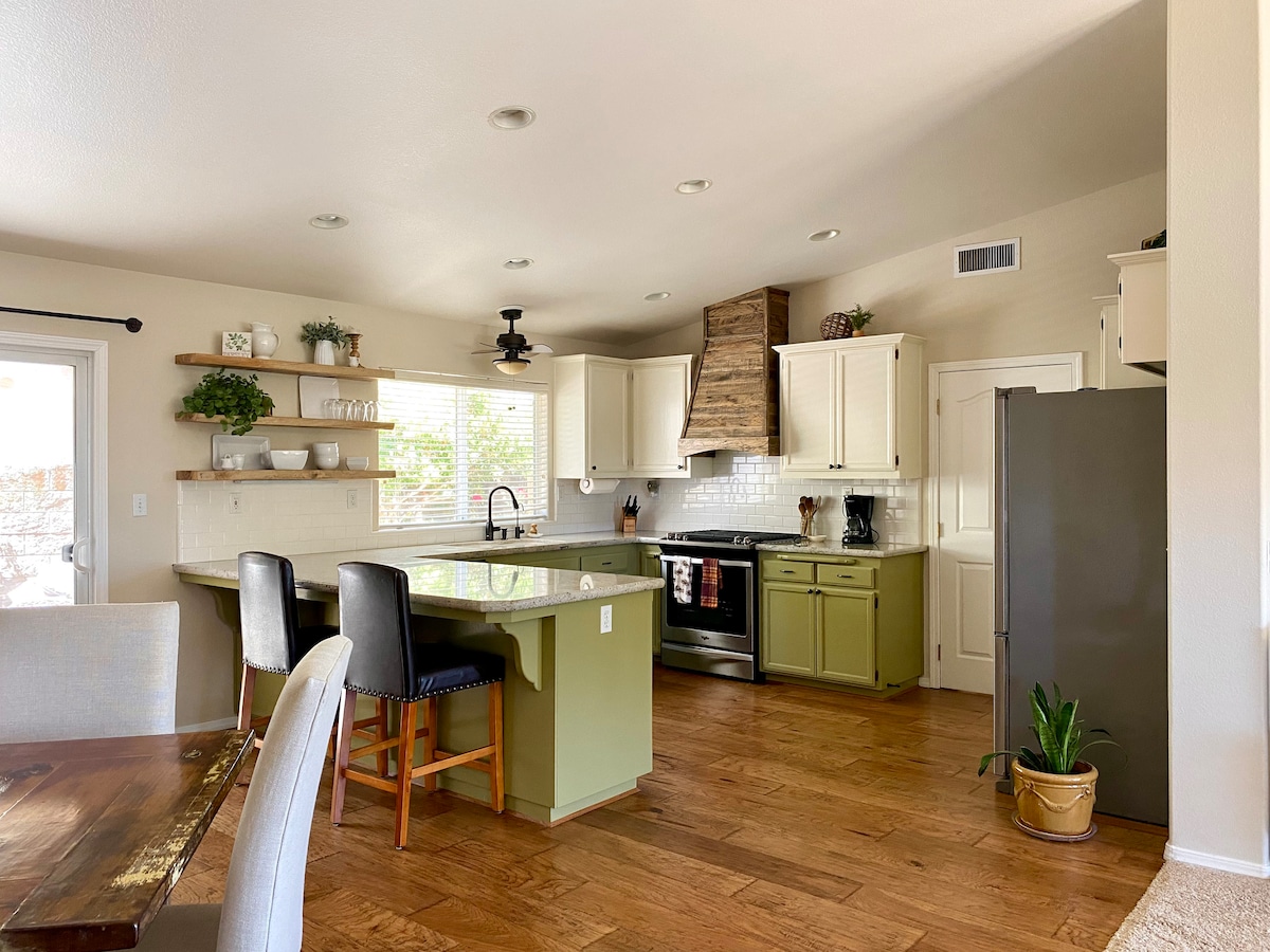 The kitchen features a spacious layout with an island and seating for three. Light cabinetry is complemented by dark countertops and a stylish backsplash. Appliances include a stove and refrigerator, while natural light streams through the windows, highlighting the wood flooring and additional shelving.
