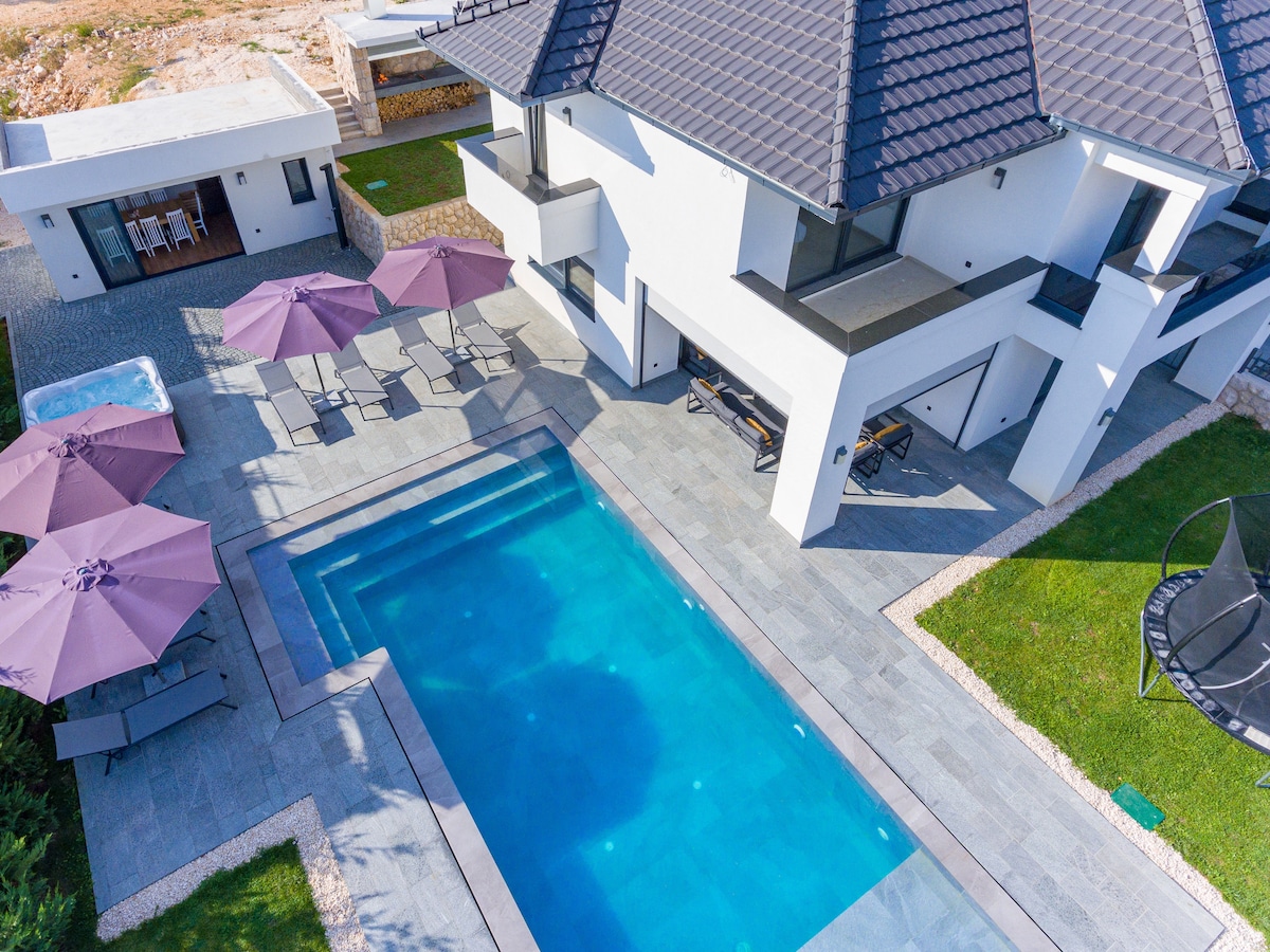 An aerial view of a modern villa featuring a rectangular swimming pool surrounded by gray stone tiles. Comfortable lounge chairs sit beneath purple umbrellas, while a hot tub is positioned nearby. The neatly landscaped yard is highlighted by green grass and stone borders.