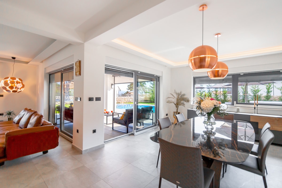 A spacious dining area features a large round table surrounded by stylish chairs. Large glass doors open to an outdoor space, connecting the indoor and outdoor environments. Copper pendant lights add a modern touch, complemented by a cozy seating arrangement in the background.