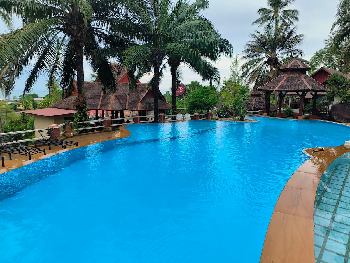 A large swimming pool with an inviting blue surface is surrounded by lush palm trees and tropical foliage. Lounge chairs line one side of the pool, while traditional style structures are visible in the background, offering shaded areas for relaxation.