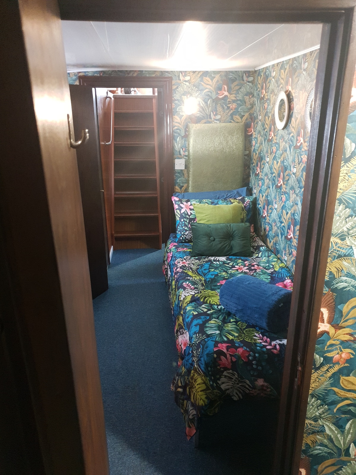 A narrow hallway leads into a cozy room, featuring vibrant floral wallpaper. A single bed adorned with colorful pillows rests against one wall, with a plush throw nearby. A wooden wardrobe stands opposite, providing storage, while a bright light illuminates the space.