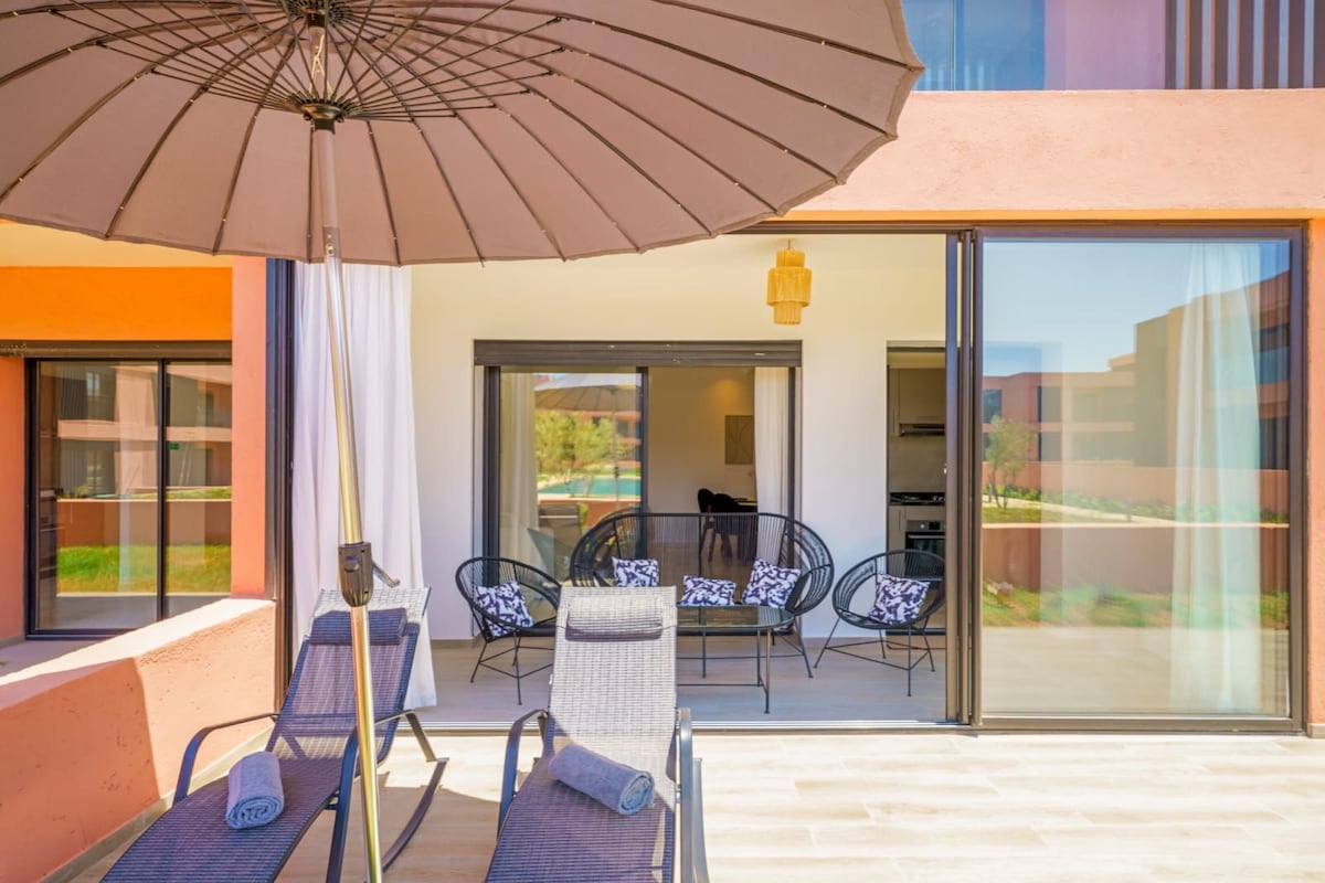 A spacious terrace area features two lounge chairs and black wicker seating, creating a comfortable outdoor space. An umbrella provides shade, while large glass doors allow natural light to enter the adjacent indoor area, showcasing a view overlooking the pool.