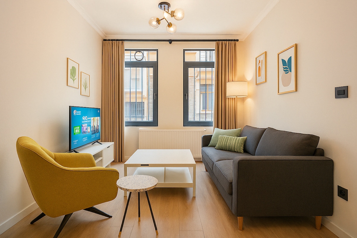 The living room features a comfortable gray sofa and a bright yellow accent chair positioned next to a white coffee table. A smart TV is visible, with natural light coming through two large windows adorned with light curtains. Art prints are hung on the walls.