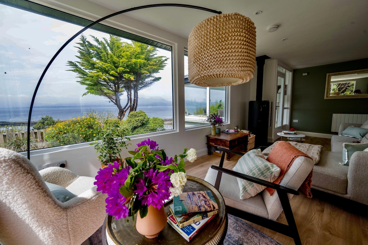 A cozy living space features large windows offering panoramic views of the ocean and surrounding greenery. A round table is topped with a vase of vibrant flowers. Comfortable seating includes an armchair and a light-colored couch. Soft lighting is provided by a woven pendant lamp.