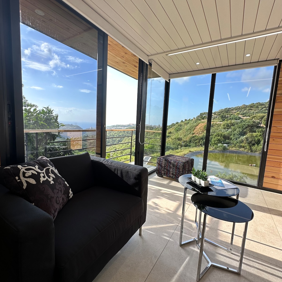 A bright seating area is enclosed by floor-to-ceiling glass walls, offering scenic views of rolling hills and a water feature. A dark sofa adorned with patterned cushions faces a small coffee table, accompanied by a vibrant pouf and a decorative plant.