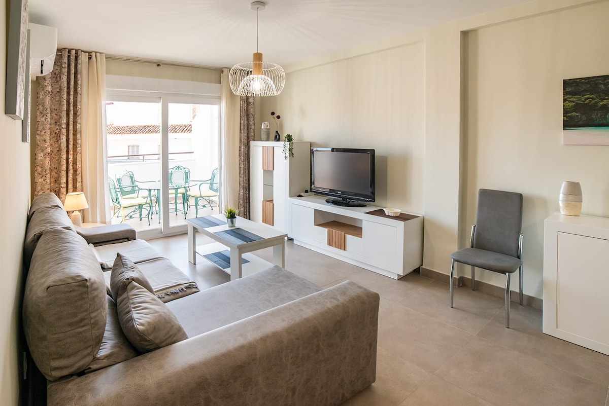 A bright living area features a comfortable sectional sofa and a sleek coffee table. A television is positioned on a white media console and natural light pours in through the sliding glass doors, which lead to a small balcony with outdoor seating.