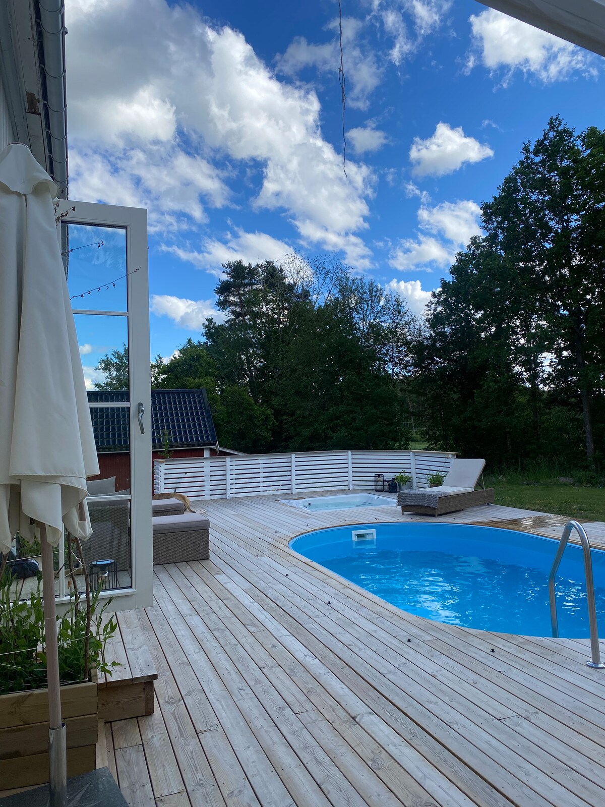 An outdoor area features a wooden deck surrounding a swimming pool, with a light blue water surface reflecting the sky. Comfortable loungers are positioned nearby, offering a space to relax. Lush greenery is visible in the background, enhancing the peaceful outdoor setting.