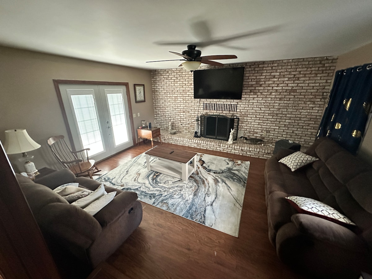 The living room features a spacious layout with two comfortable couches and a rocking chair. A large flat-screen TV is mounted above a brick fireplace. Light-colored curtains frame double glass doors, while a coffee table sits atop a patterned rug that enhances the warmth of the wooden floor.