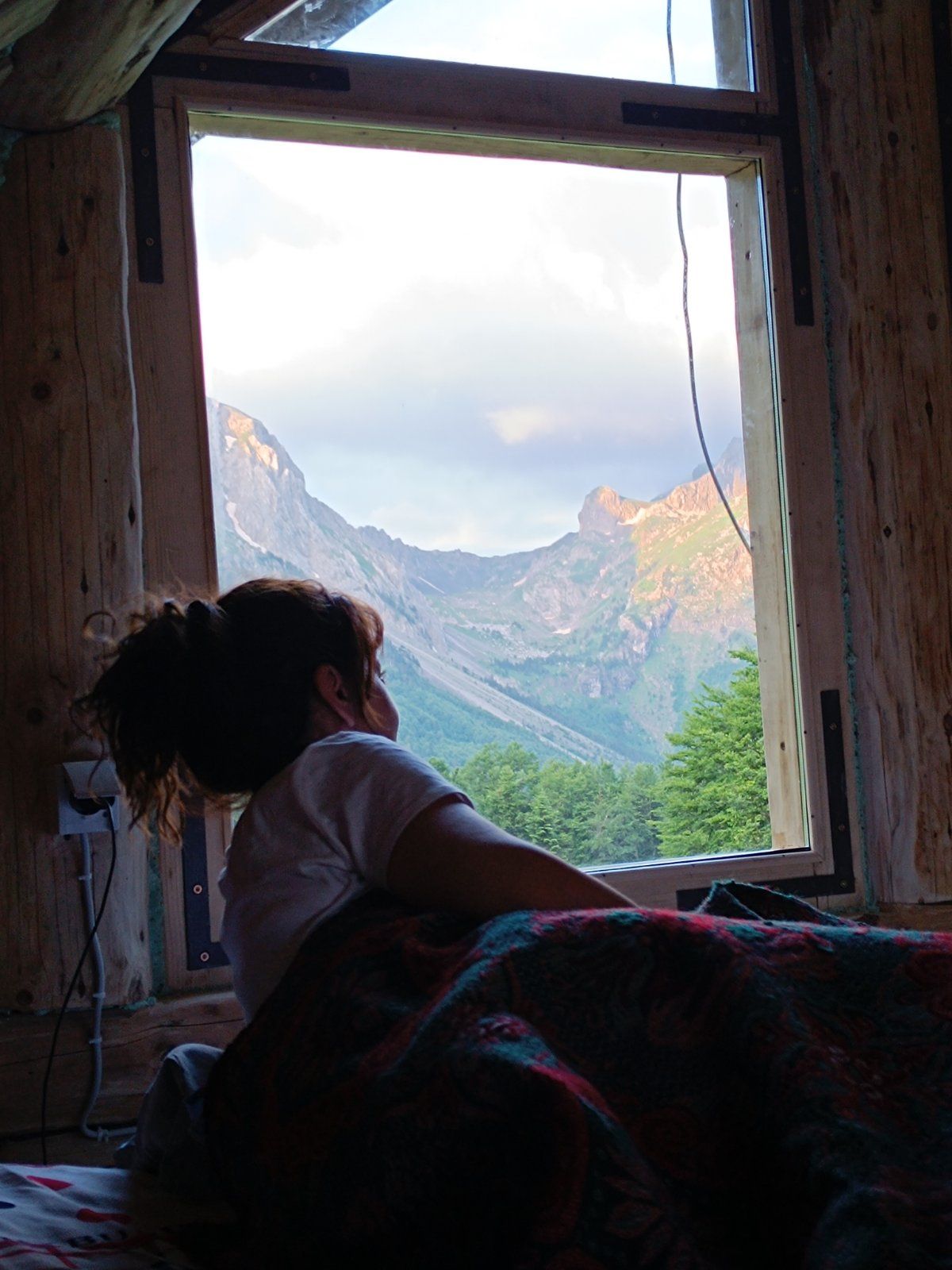 A window reveals a stunning mountain landscape, featuring rugged peaks and greenery below. Natural light filters into the room, casting soft shadows. A guest is seated on a bed, gazing thoughtfully at the view while wrapped in a colorful blanket.