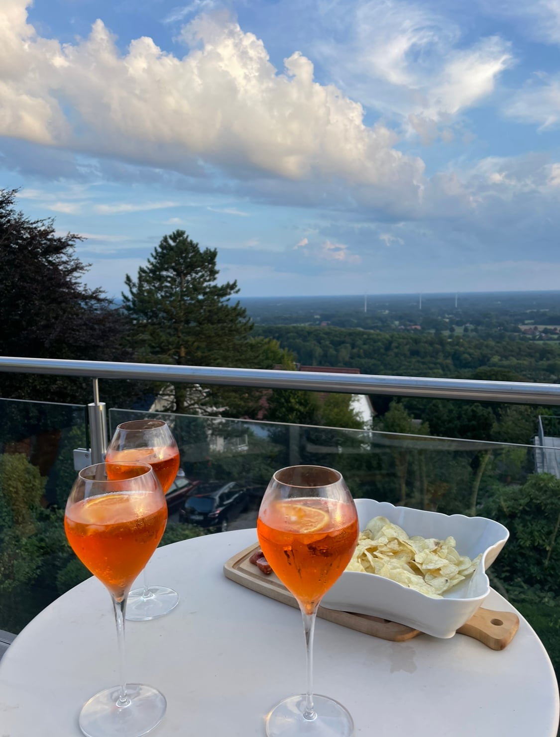 Three glasses of orange beverages are set on a round table. A bowl of chips is placed beside the drinks. The scene overlooks a lush landscape, with distant hills and a cloudy sky in the background, creating a serene outdoor setting.