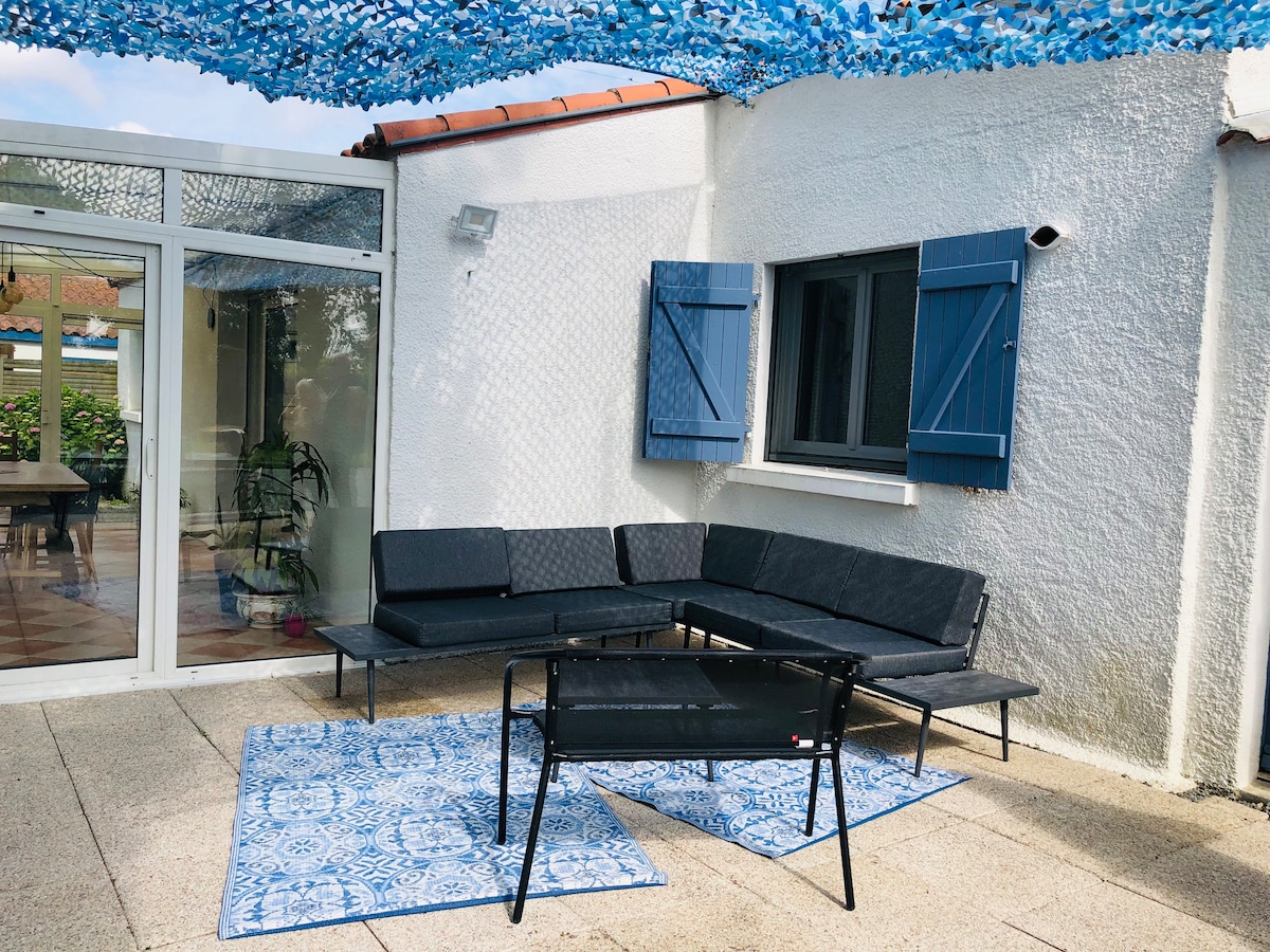 An outdoor terrace is furnished with a corner sofa in dark tones and a black chair. A patterned blue rug is placed on the floor, complemented by blue-patterned fabric overhead. Light filters through open blue shutters of the adjacent room, creating a calm atmosphere.