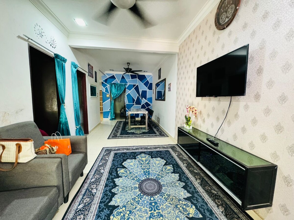 A spacious living area is displayed, featuring a large flat-screen TV mounted on the wall and a contemporary glass coffee table in the center. The room is adorned with ornate rugs and accentuated by teal curtains, providing a vibrant ambiance.