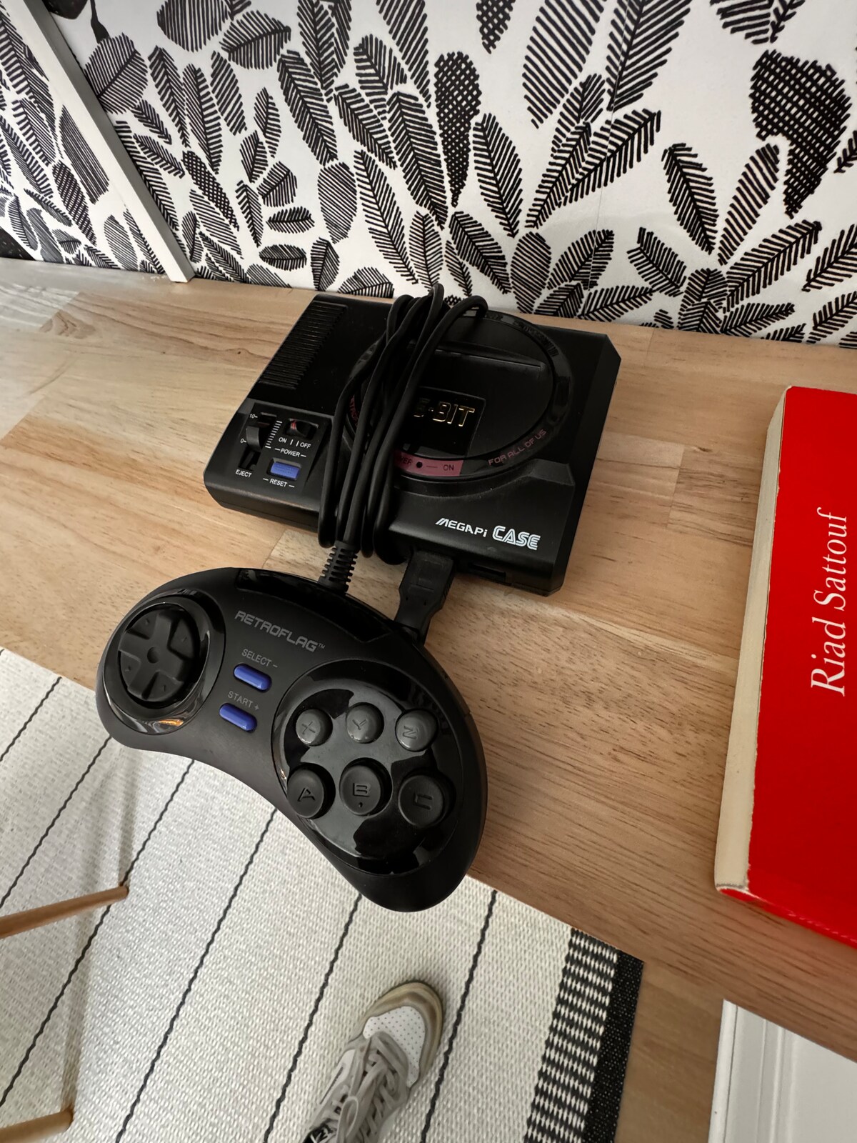 A classic gaming console is displayed on a wooden surface, with a wired controller resting adjacent to it. The backdrop features a stylized black and white patterned wallpaper, adding a modern touch to the vintage gaming setup.