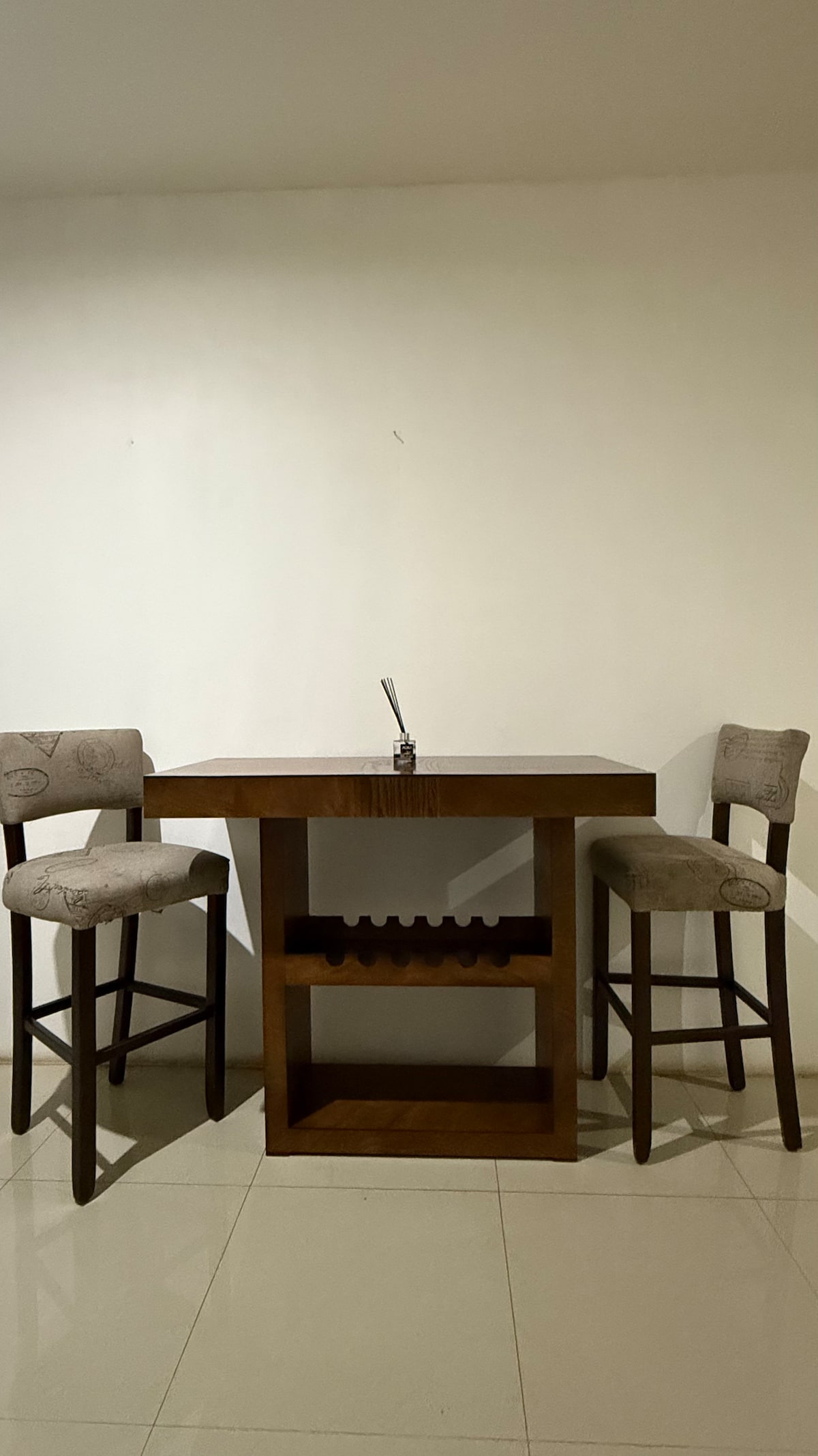 A small dining area features a compact wooden table accompanied by two upholstered bar stools. The table is positioned against a plain white wall, with a minimalist decorative piece placed centrally on top. A small shelf is visible beneath the tabletop.