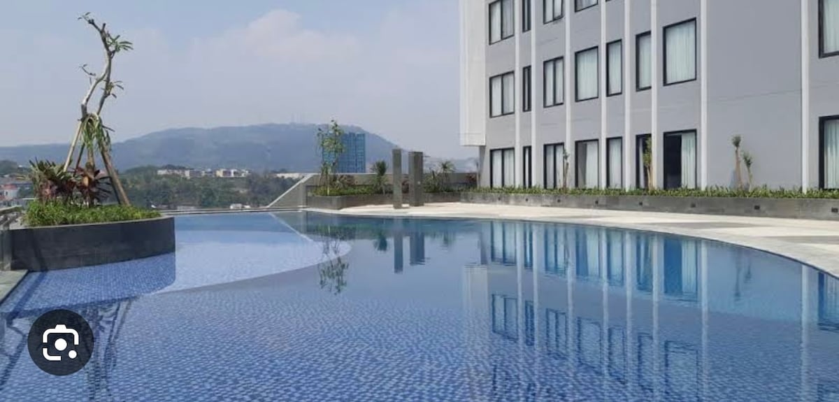 A serene swimming pool reflects the clear sky, bordered by a modern building. Lush greenery and decorative plants enhance the space, while distant hills provide a scenic backdrop. The pool's surface features shimmering blue tiles, creating an inviting atmosphere for relaxation.