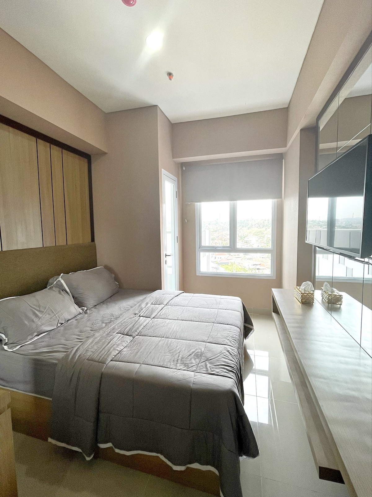 A well-appointed bedroom features a spacious bed with neutral-toned bedding. A flat-screen TV is mounted on the wall opposite the bed, while a large window allows natural light to fill the room. Light-colored walls contribute to a calm and inviting ambiance.