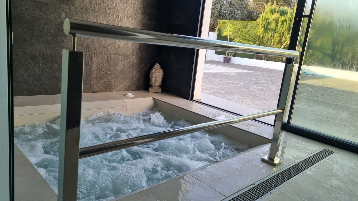 A jacuzzi is featured in a sleek, modern bathroom, surrounded by minimalist design elements. The water is bubbling, creating a calming effect. A safety railing provides support, while a large glass window offers a view of the outdoor area, enhancing the serene setting.
