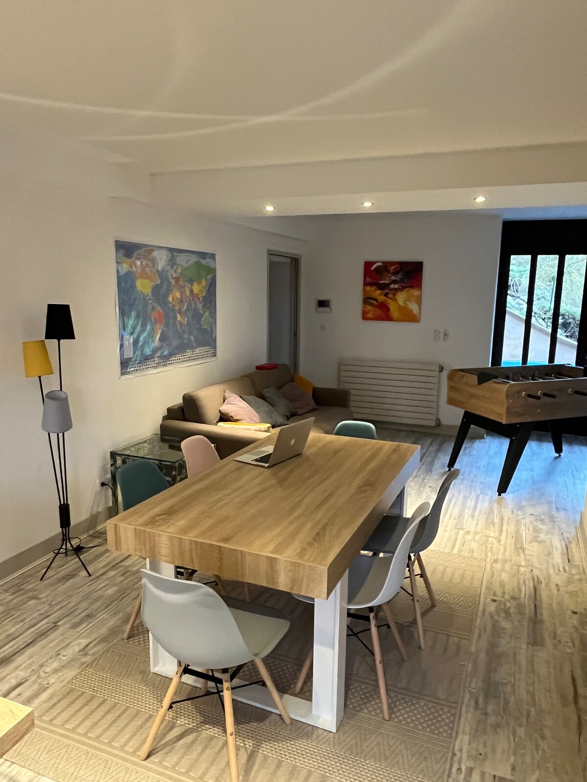 A modern dining area features a large wooden table surrounded by several chairs in varied colors. A comfortable seating space is arranged nearby with a sofa and colorful wall art, while a foosball table is positioned in the corner, enhancing the communal atmosphere.