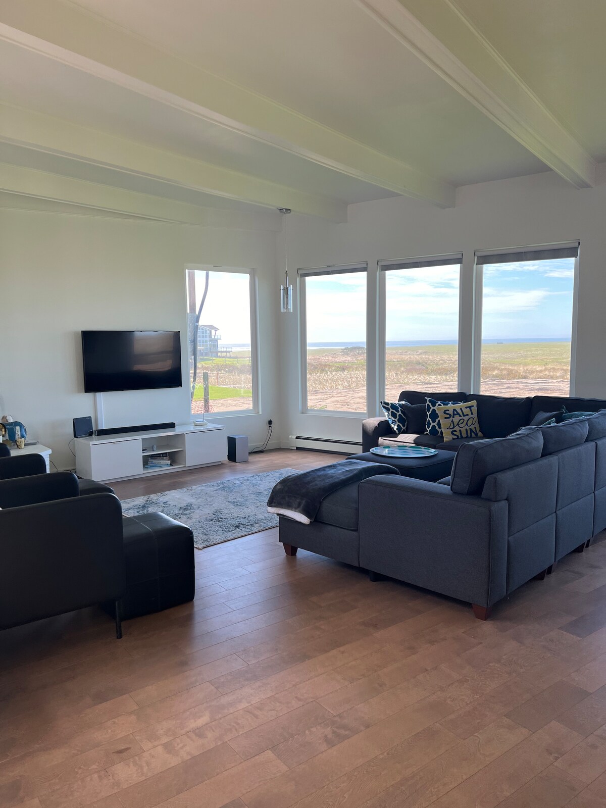 A bright living room is presented with large windows showcasing views of the outdoors. Modern furniture is arranged around a central area, featuring a television mounted on the wall and a soft rug covering the wooden floor.
