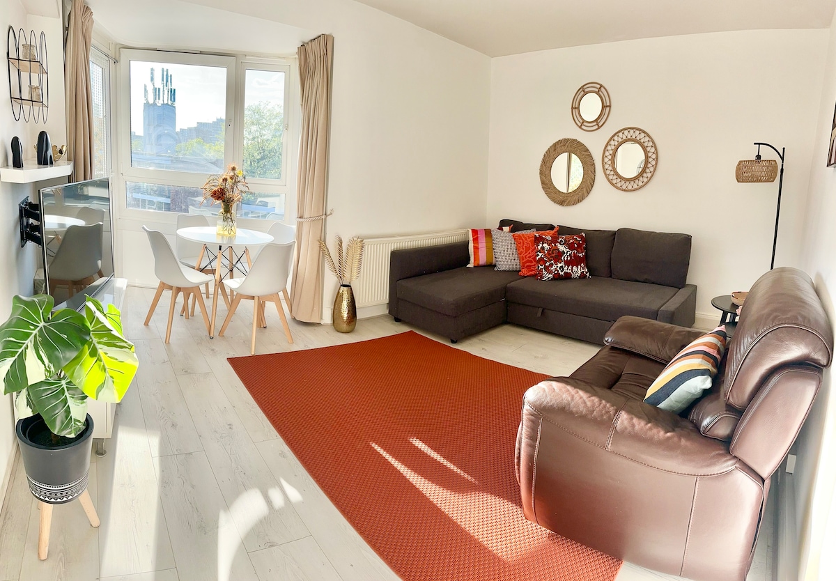 The living room features a comfortable sectional sofa and an armchair, arranged around a warm-toned area rug. A dining table with four chairs is positioned adjacent to the large windows, allowing natural light to fill the space. Decorative mirrors adorn the walls.