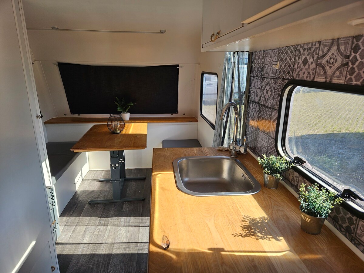 An intimate dining area features a wooden table paired with a bench, illuminated by natural light. A compact kitchen is equipped with a sink and decorative potted plants, showcasing a combination of modern design and natural elements. The walls exhibit stylish patterned details.