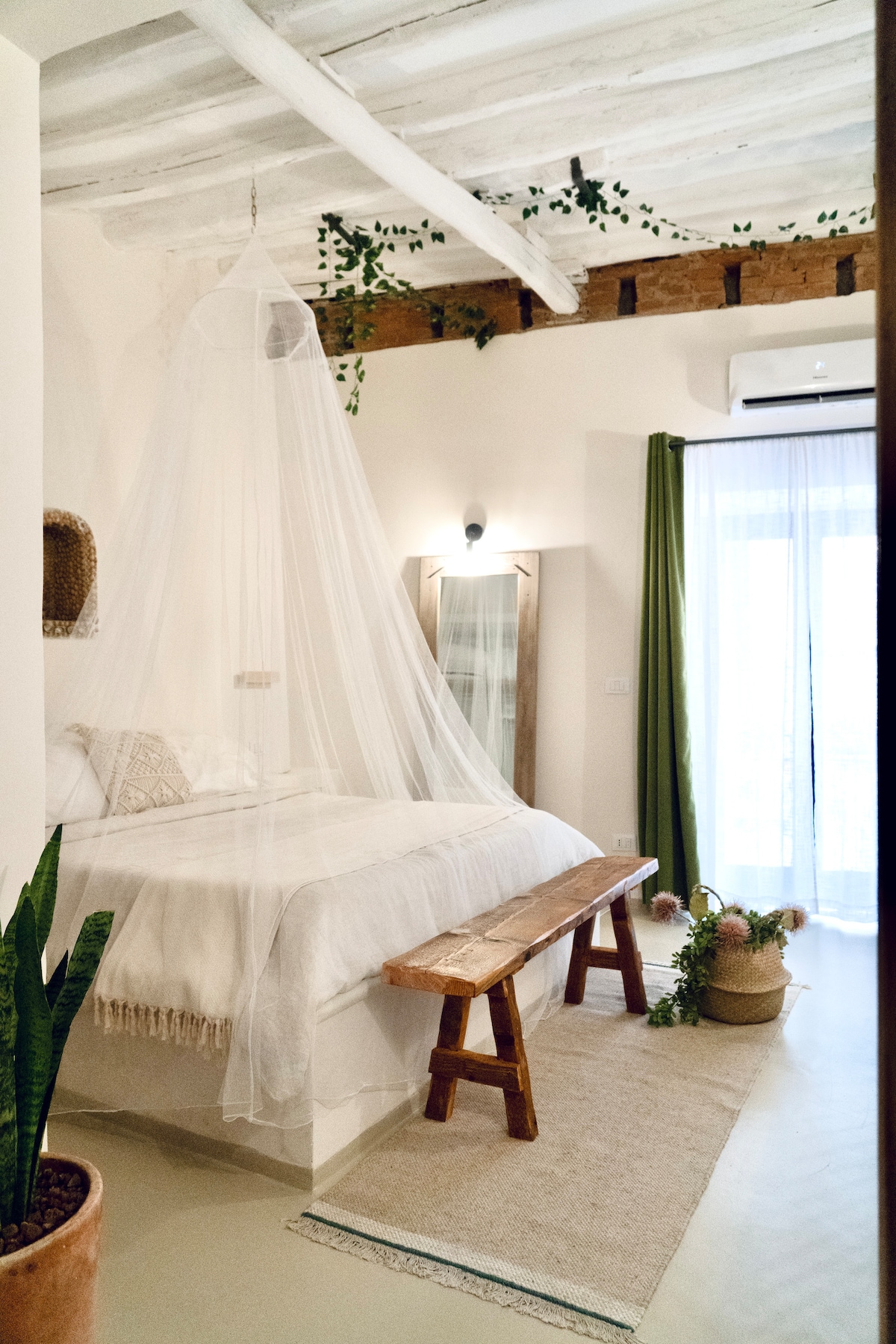 The bedroom features a spacious bed draped with a light canopy, enhancing the sense of tranquility. A wooden bench provides additional seating. Natural greenery decorates the space, while soft curtains frame the balcony doors, allowing light to filter in.
