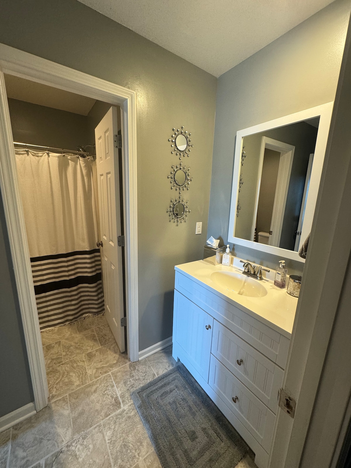 The bathroom features a modern vanity with a countertop and a mirror. A shower area with a striped curtain is visible, while light-colored tiles cover the floor. Decorative mirrors hang on the wall, enhancing the space's contemporary feel.