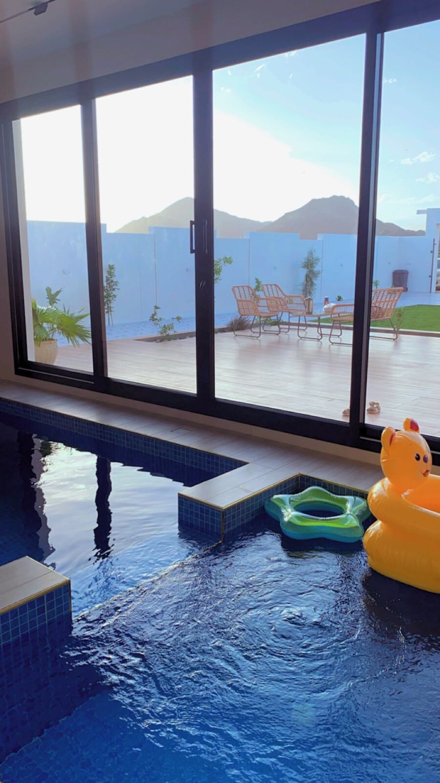 An indoor swimming pool is visible, featuring still water and a yellow inflatable ring. Large glass doors open to a deck with outdoor seating and a green lawn, framed by distant mountains. Potted plants are placed near the pool area.