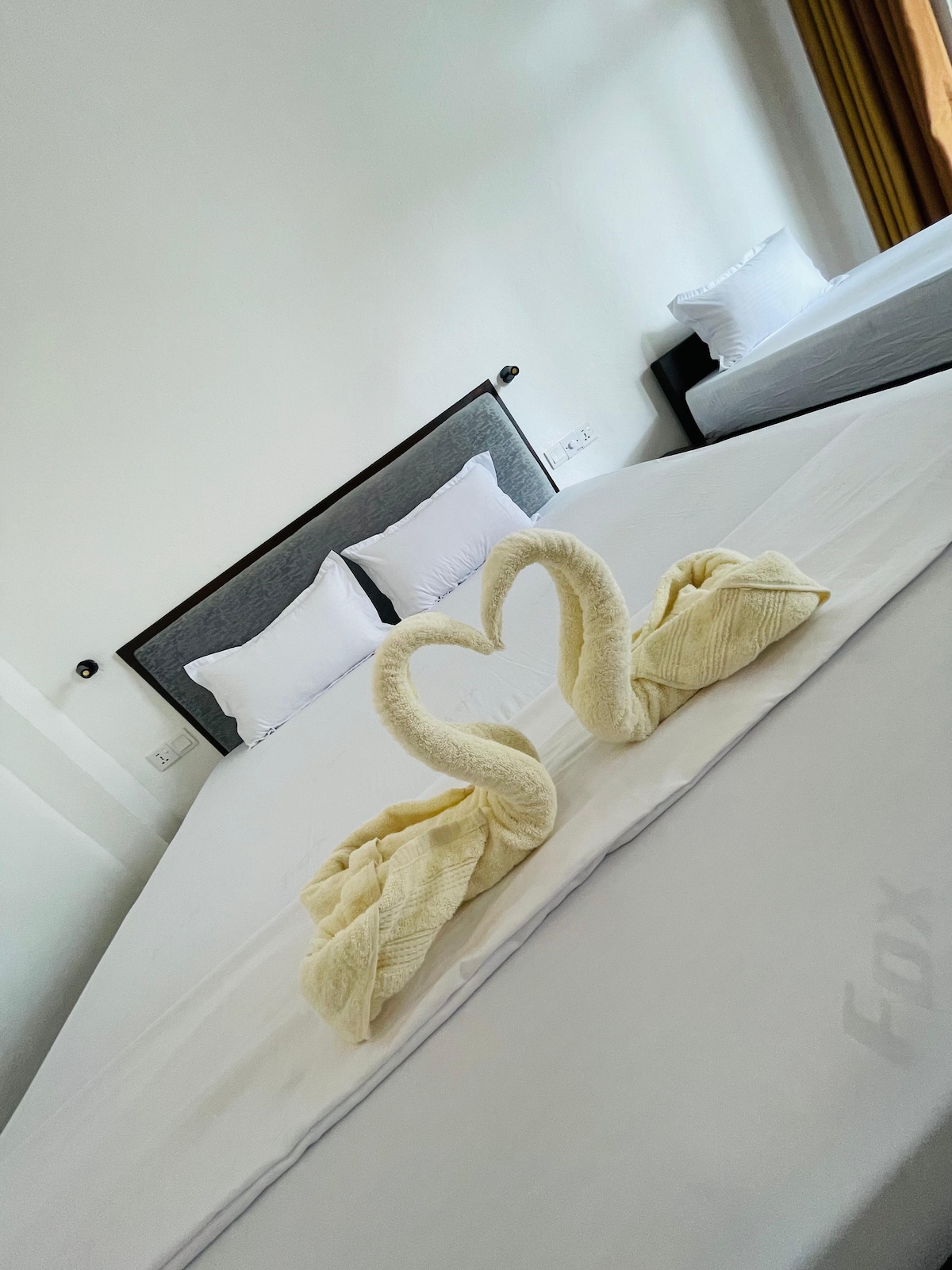 A spacious room features two beds with neutral-toned bedding. Decorative towel swans are positioned in a heart shape at the center of the larger bed. Soft lighting enhances the serene atmosphere. Large windows with light curtains allow natural light to fill the space.