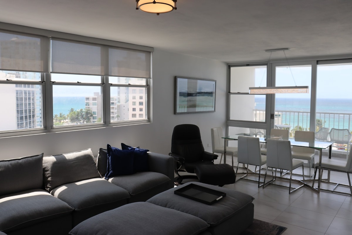 A spacious living area is captured, featuring a gray sectional sofa and a black lounge chair. Large windows allow for ample natural light and showcase views of the ocean and palm trees. A dining table with six chairs is positioned nearby, creating an inviting atmosphere.