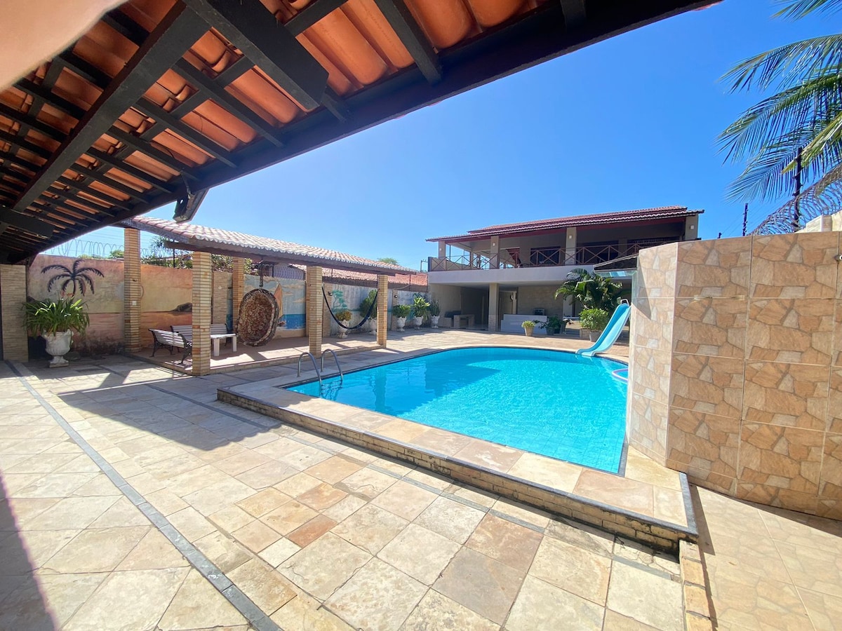 A spacious outdoor area features a vibrant blue pool surrounded by stone tiles. A slide is visible at one end, while shaded seating areas are provided under the roof. Palm trees and a waterfall accentuate the tranquil environment.