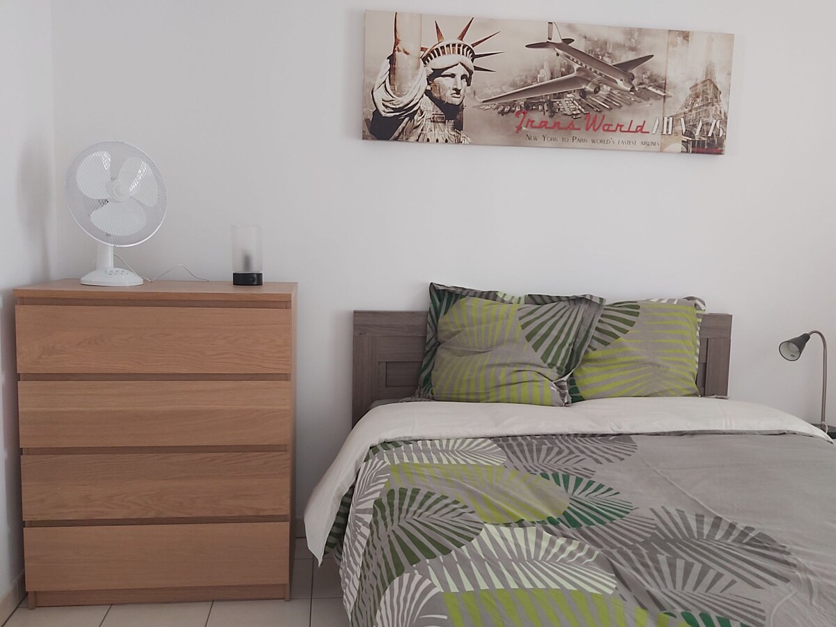 A spacious sleeping area is displayed, featuring a neatly made bed with patterned bedding and a colorful accent. A wooden dresser with three drawers is positioned nearby, accompanied by a small lamp and a desk fan on top. Wall art adds character to the room.